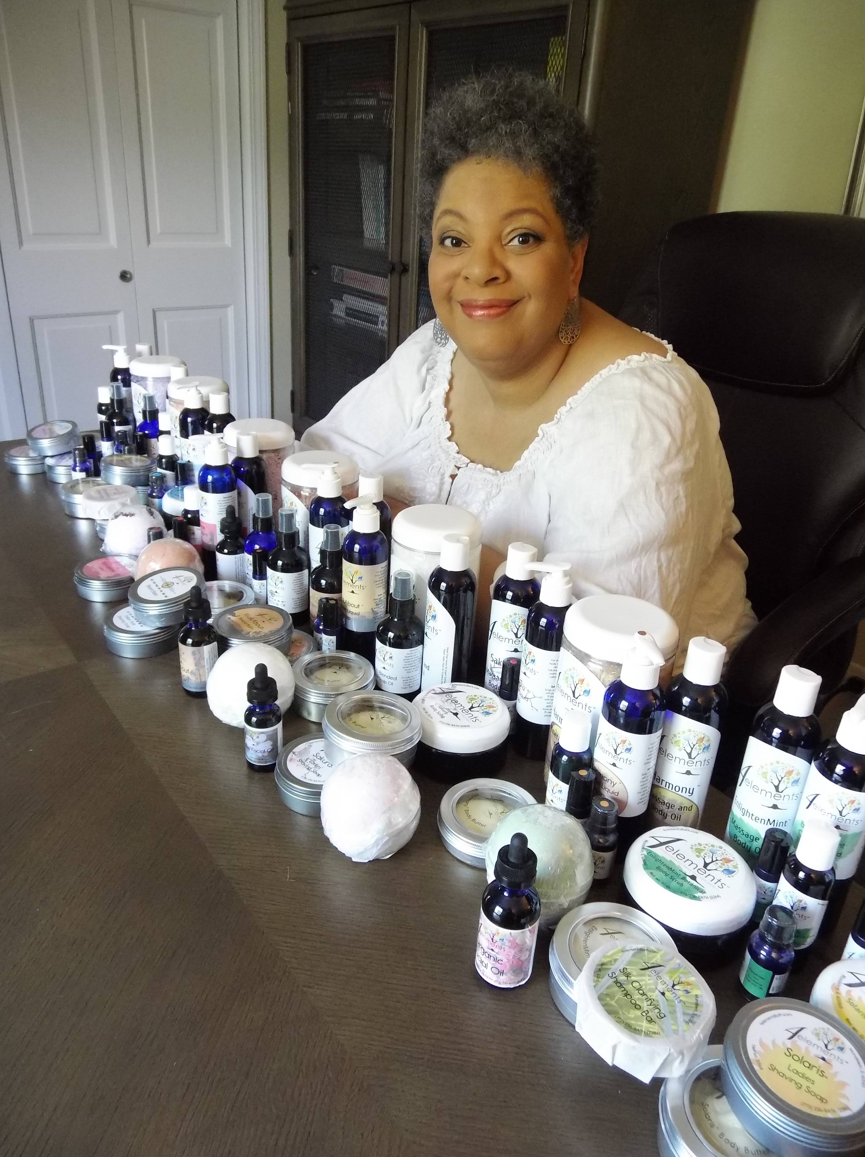 4Elements Bath Founder Charise Cowan-Leroy smiling behind a spread of skincare products such as lotions, body butters, and castile soaps.