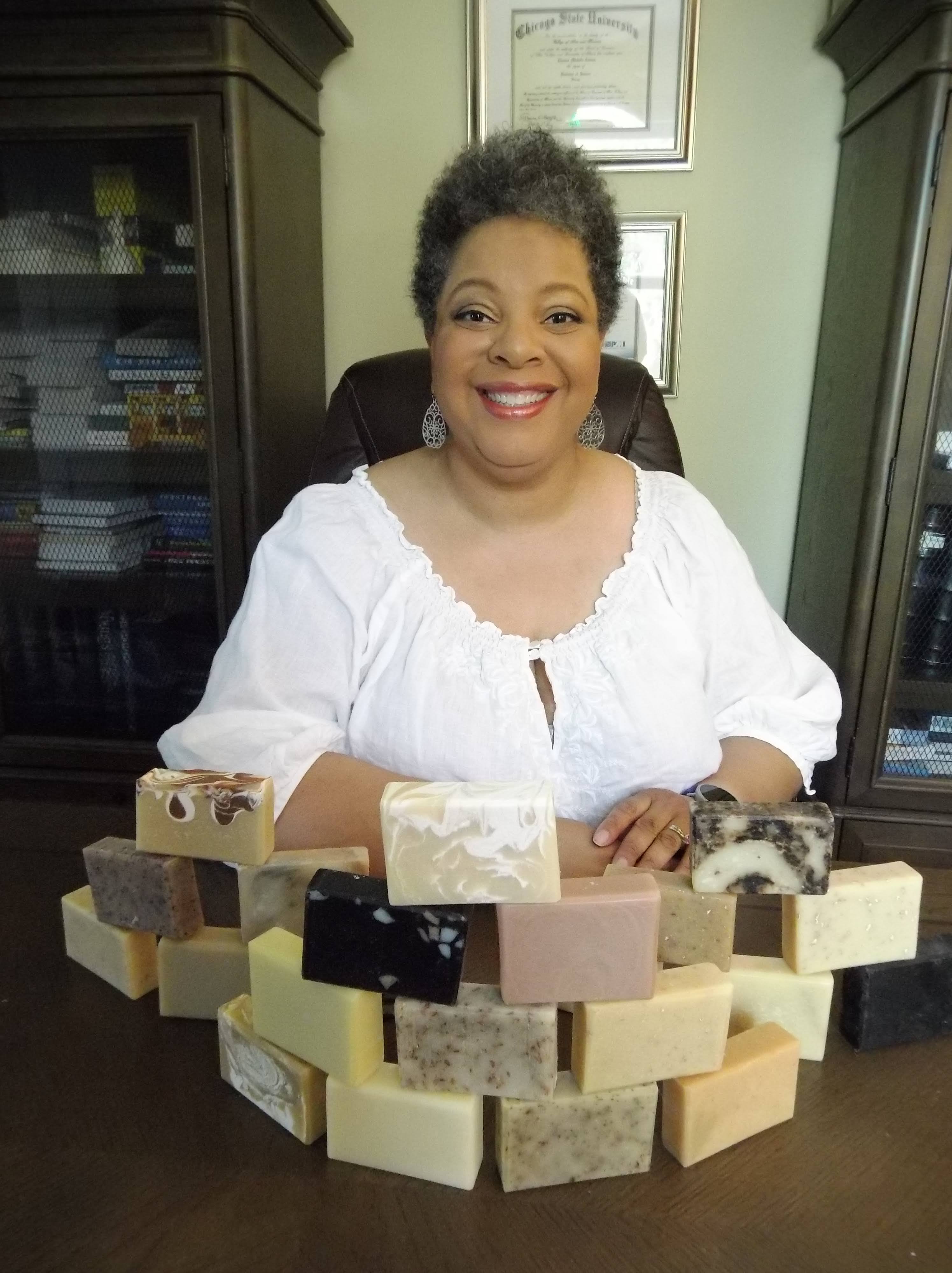 4Elements Founder Charise Cowan-Leroy in a white shirt smiling behind a stacked display of brown, white, and charcoal soaps.