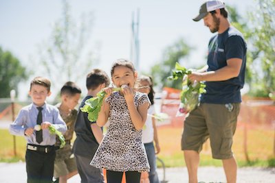 young girl eating radish with other students and teacher behind her in the climate victory garden