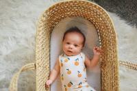 baby laying in crib wearing a onesie with oranges on it, looking curiously at the camera. The crib is wicker and is on top of a white faux fur rug.
