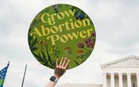 A white hand holding a round, green sign that reads: "Grow Abortion Power" in front of the Supreme Court building