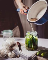A hand holding a blue pot pouring liquid into a mason jar full of pickling cucumbers.