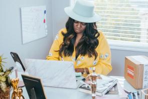 A Black woman sits at a desk in front of a laptop. She's wearing a wide-brimmed white hate and yellow floral blouse, she's looking down at the laptop. On the desk are various items - a picture frame, a box, a statue, a notebook. On the wall next to her is a whiteboard with scribbles on it and behind her is a window. Black-Owned Businesses for Black History Month. Juneteenth.