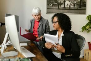 A Black woman sits at a desk in front of a computer desktop and keyboard with a clipboard in her hands. An older white woman in a grey blazer stands beside her with a folder of papers. Tax Planning Sustainable Growth Business.