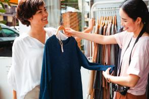 Two Asian women shopping outside, photo is from the hips up. Woman on the right wears a pale pink shirt with a camera on a strap around her neck. She holds up a dark blue sweater on a hanger with a tag in front of the other woman with short hair wearing a white blouse. Trust small businesses.