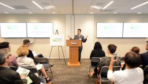 This picture features, Adam Roberts, executive director of Bethesda Green, standing at a podium in the front of a room filled with people. Two  large screens on either side of him, with the text "Bethesda Green Summer Pitch Event" 