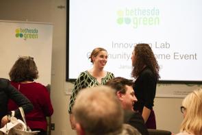 This photo features two women in discussion, smiling at one another in front of a large screen that reads "Bethesda Green, Innovation Lab Community Welcome Event"