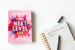 A photo of a pink book with a water color like splash of color titled Your Next Level Life. Book is next to an open spiral notebook with some writing and a pen resting on top.