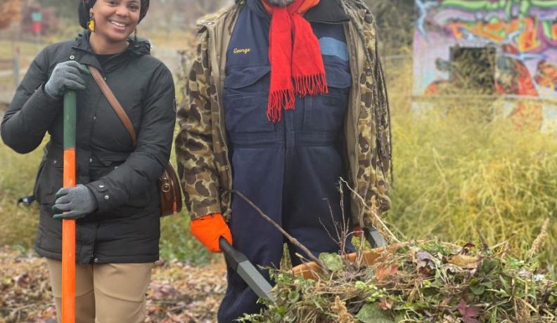 Michael Chaney is wearing a big brown coat and a red scarf, holding a wheelbarrow filled with foliage for compost. He stands next to Jasmira Colon who is smiling at the camera and holding a shovel with an orange handle.