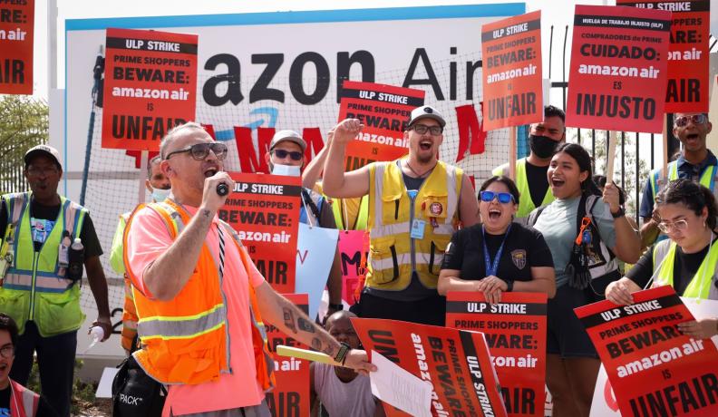 a group of Amazon workers striking. They are holding up red signs and yelling together.