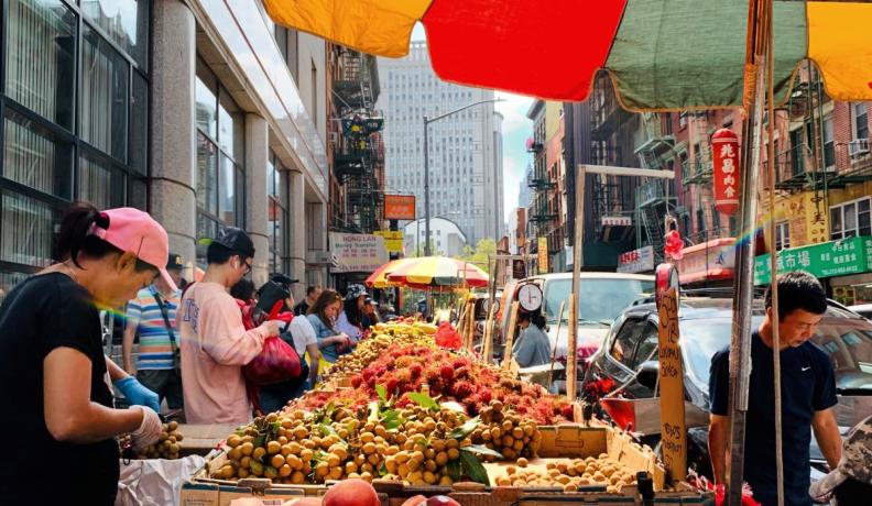 a fruit market in NYC. Close up is a stand of nectarines under a red, yellow, and blue sun parasol. In the background are more fruit stands and onlookers.