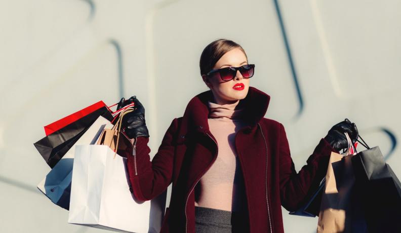 A white woman is dressed very chic in a red coat and dark sunglasses. She is holding numerous paper shopping bags in both hands like she just finished a big shopping spree.