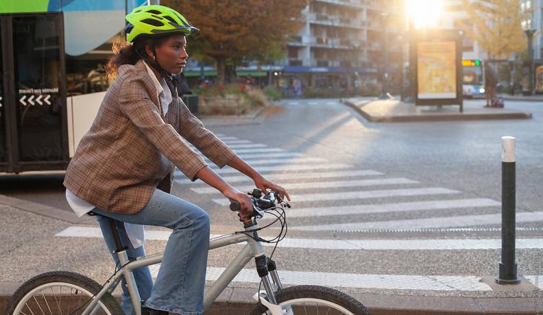 Black woman in a brown blazer, flared jeans, and a neon green helmet riding a gray bike through city streets.
