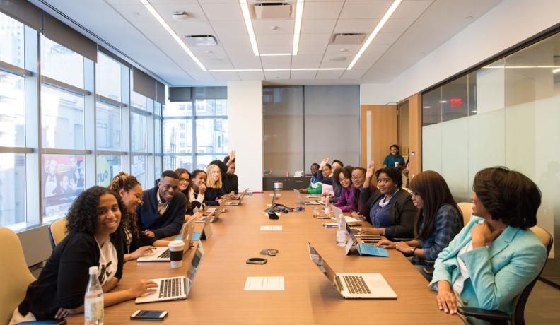 A look at a conference table from the head of the table. Over 16 people are talking among each other and themselves, with laptops in front of them, and three are raising their hands.
