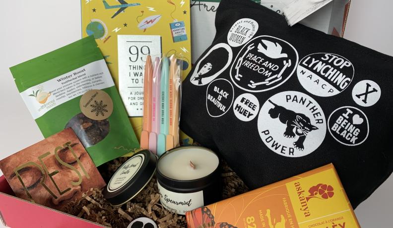 A box filled with various goodies, including pens, a candle, a book, chocolate, and more.