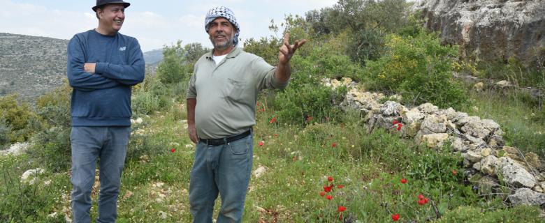Nasser Abufarha stands to the left on a grassy hill in Palestine, arms crossed in a long-sleeve blue shirt and black hate. On the right is a local Palestinian farmer, in a grey shirt and turban, as they discuss regenerative agriculture.