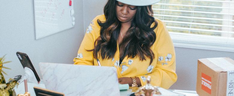 A Black woman sits at a desk in front of a laptop. She's wearing a wide-brimmed white hate and yellow floral blouse, she's looking down at the laptop. On the desk are various items - a picture frame, a box, a statue, a notebook. On the wall next to her is a whiteboard with scribbles on it and behind her is a window. Black-Owned Businesses for Black History Month. Juneteenth.