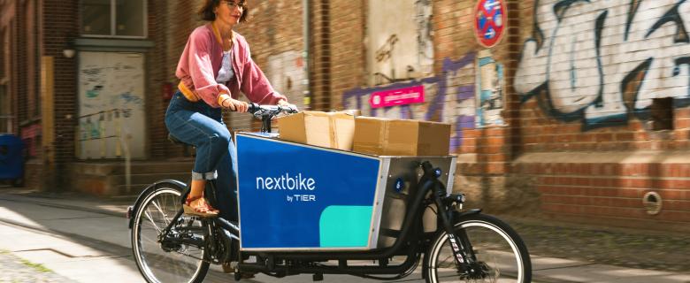A white woman with a short brunette bob wearing a pink sweater and jeans rides a bike in front of a brick wall with graffiti. The bike has a big plastic bucket between the two wheels for transporting packages. Green Businesses in 2023.