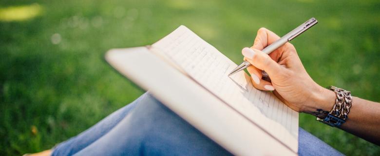 A woman's hand writes in a notebook resting on her knees. She's sitting in a grass field. Sustainable creative accessories.