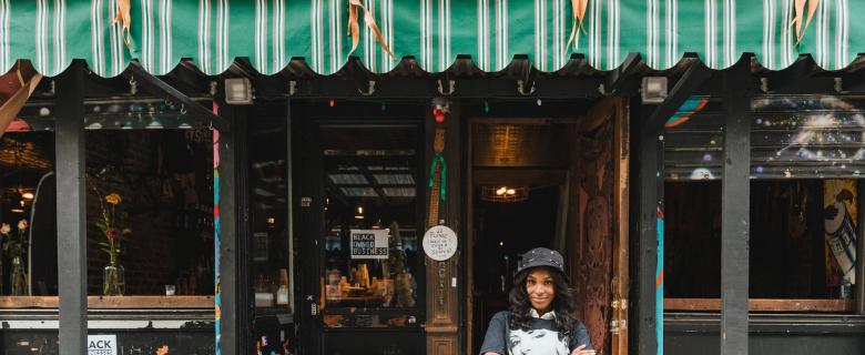 A Black woman business owner stands outside her establishment. Green business resolutions.