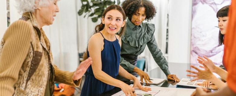 A group of women stand around a table covered in paper. They are actively discussing something. Women Business Owners.