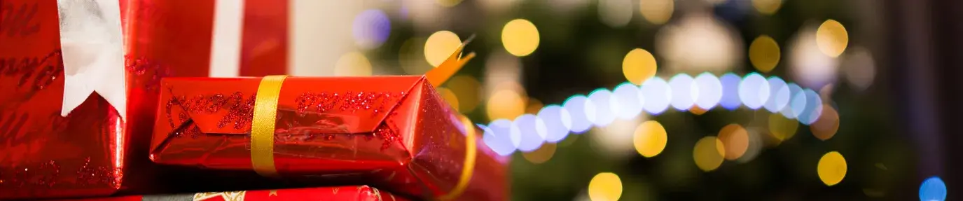 a stack of three presents wrapped in red, shiny paper are sitting in front a Christmas covered in yellow and blue lights.
