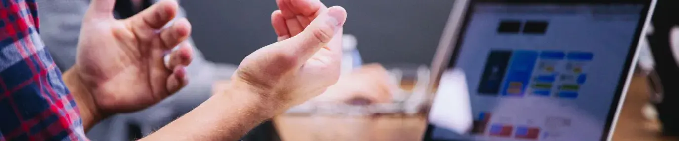 hands gesturing to explain something in a meeting. Computer and notebook are laid out on a table.
