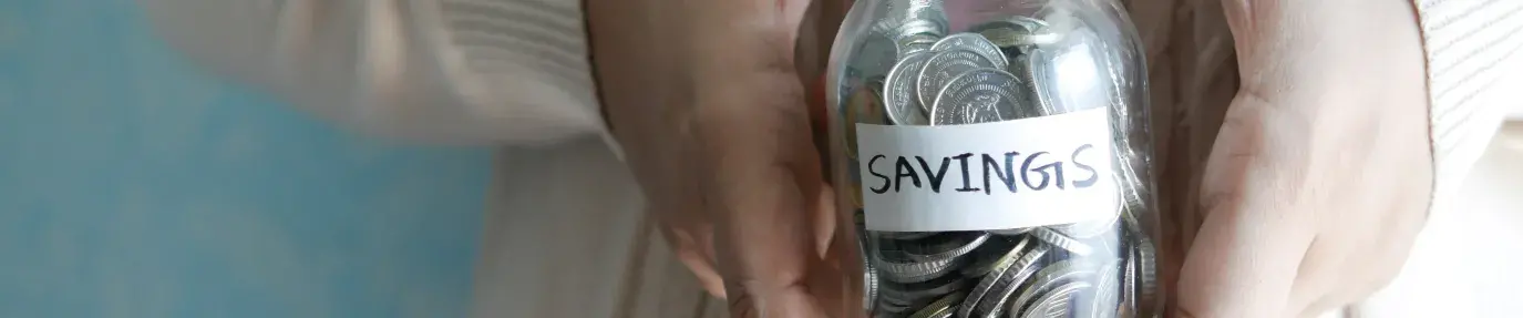 A pair of hands holding a small mason jar filled with coins and a label on the jar that reads: "Savings"