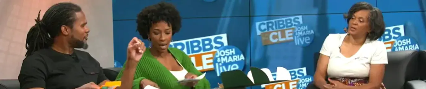 Gloria Ware, a Black woman, sits on the far right, being interviewed at a local TV news station. On the left, a Black man and Black Woman interview her.