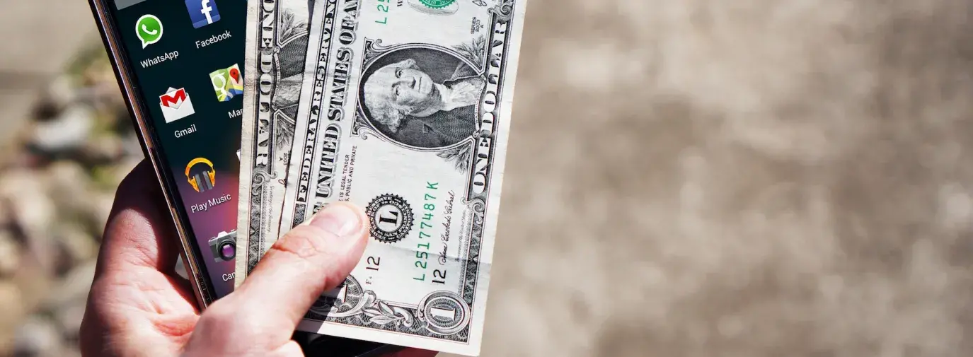 Image: hand holding smartphone and cash. Topic: What Does it Mean to Vote With Your Dollar?