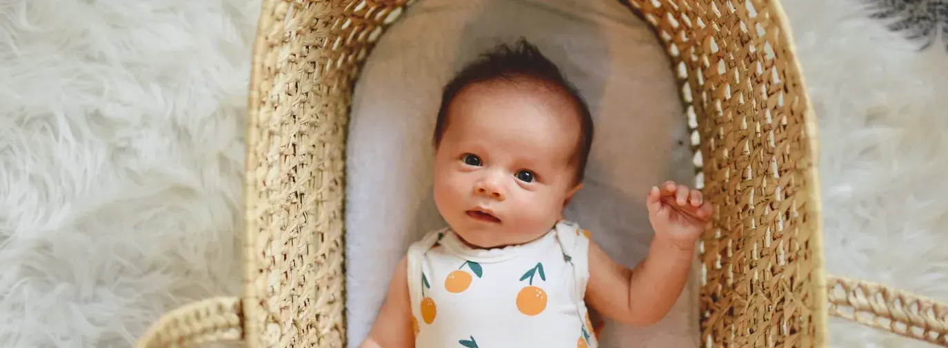 baby laying in crib wearing a onesie with oranges on it, looking curiously at the camera. The crib is wicker and is on top of a white faux fur rug. The mattress still matters for everyone.