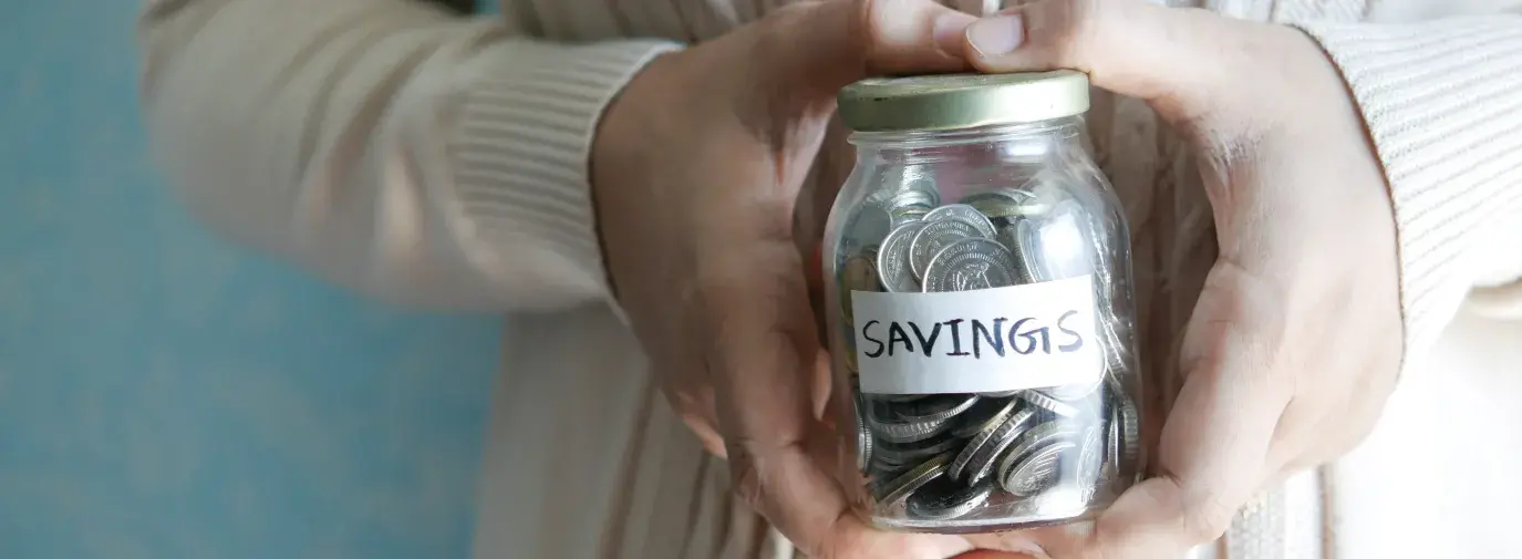 A pair of hands holding a small mason jar filled with coins and a label on the jar that reads: "Savings"