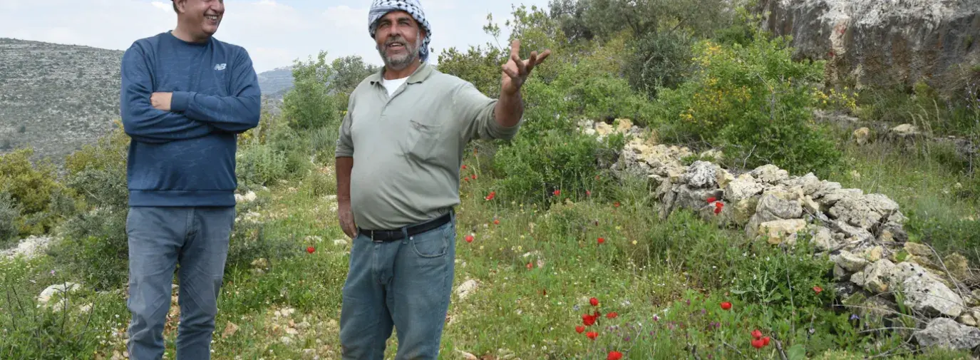 Nasser Abufarha stands to the left on a grassy hill in Palestine, arms crossed in a long-sleeve blue shirt and black hate. On the right is a local Palestinian farmer, in a grey shirt and turban, as they discuss regenerative agriculture.