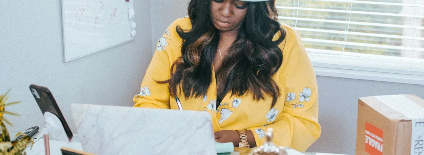 A Black woman sits at a desk in front of a laptop. She's wearing a wide-brimmed white hate and yellow floral blouse, she's looking down at the laptop. On the desk are various items - a picture frame, a box, a statue, a notebook. On the wall next to her is a whiteboard with scribbles on it and behind her is a window. Black-Owned Businesses for Black History Month. Juneteenth.