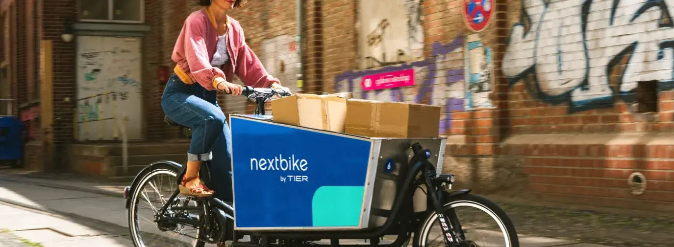 A white woman with a short brunette bob wearing a pink sweater and jeans rides a bike in front of a brick wall with graffiti. The bike has a big plastic bucket between the two wheels for transporting packages. Green Businesses in 2023.