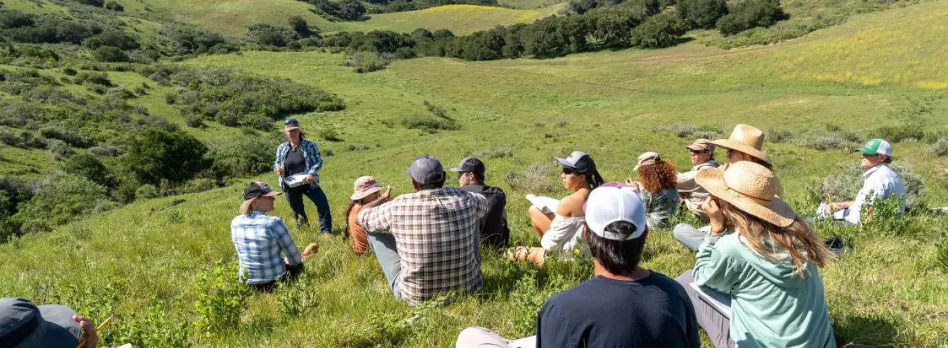 Jackie Eshelman teaching a 5-Day Ecological Outcome Verification™ course at the Center for Regenerative Agriculture at Jalama Canyon Ranch. There are people sitting on the grass all around listening to her speak with rolling green hills in the background.