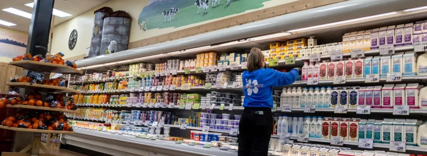A woman in a blue trader joe's employee shirt organizing the shelves in the dairy section.