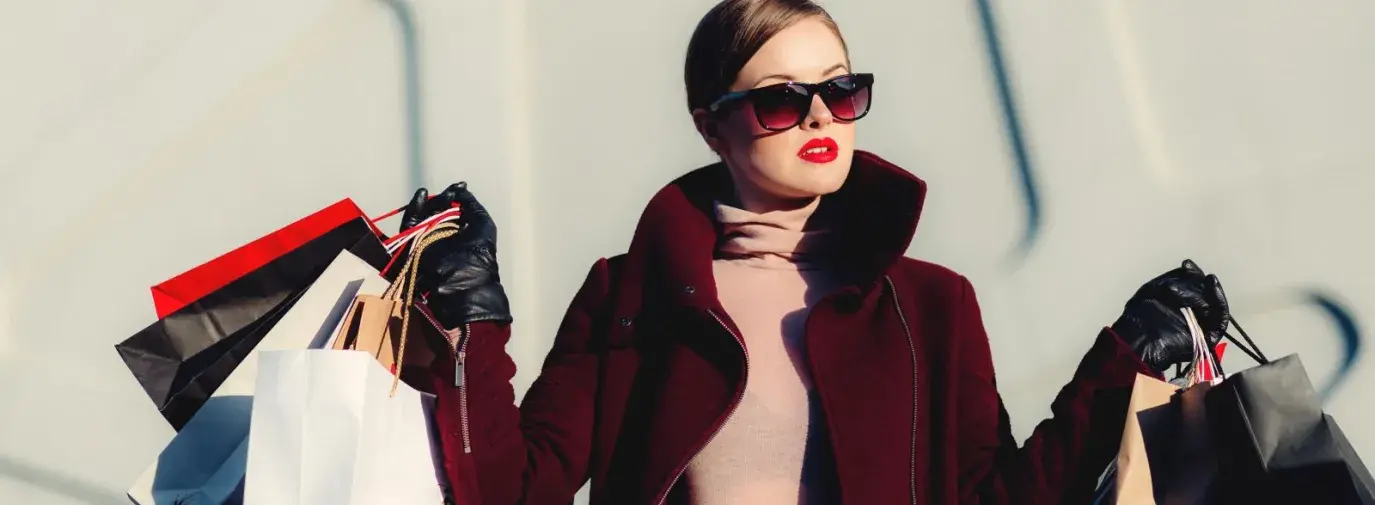 A white woman is dressed very chic in a red coat and dark sunglasses. She is holding numerous paper shopping bags in both hands like she just finished a big shopping spree.
