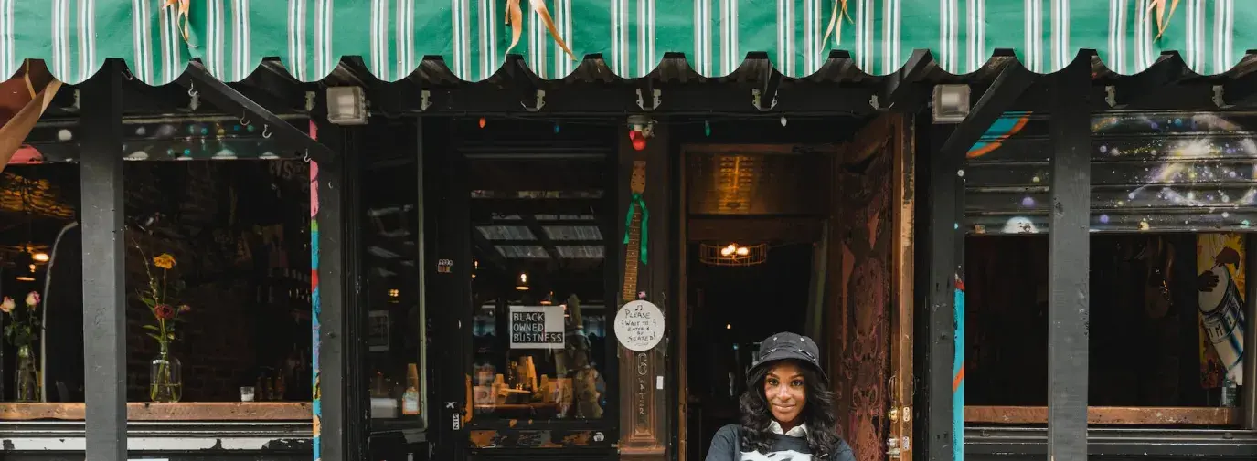 A Black woman business owner stands outside her establishment. Green business resolutions.