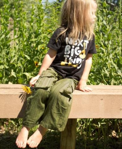 Child sitting on a wooden bench in a black t-shirt and green pants. Holds a daisy in one hand. Fair Trade Gift Guide.