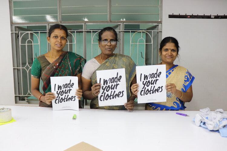 Three Indian women stand in front of a table holding signs that read: "I made your clothes." The Good Tee.