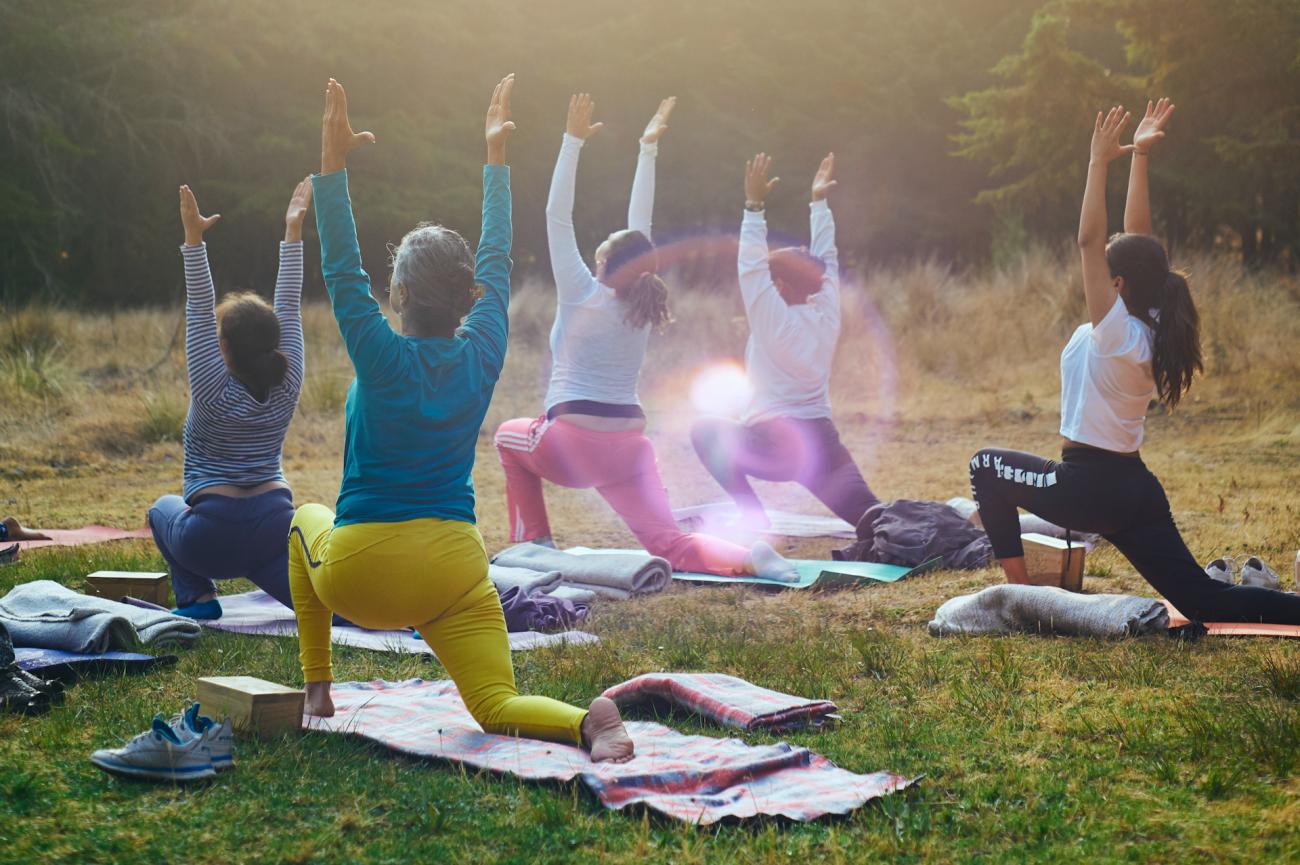 A group of people doing yoga - a forward lunge with their arms straight up - outside on grass. There is a lens flare in the middle of the photo. Mental Health.