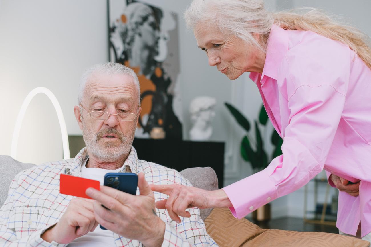 An older white man with glasses sits looking at his phone with a credit card in his hand. To the right, an older white woman in a pink button-down leans over the man, pointing at the phone screen.