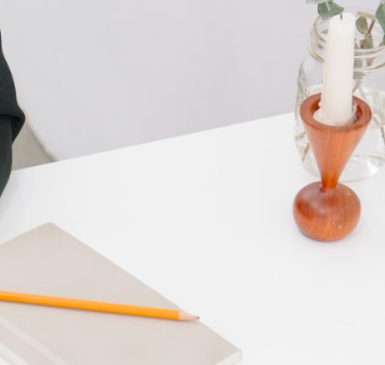 Desktop with a candle, notebook, pencil, plant. There is the arm of a person resting casually on the desk