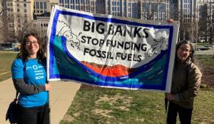 Two women are holding a sign that reads "Big Banks, Stop Funding Fossil Fuels". They are both smiling at the camera and standing in a park.