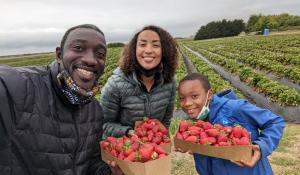 The Nortey family, made up of a man, woman, and son, are smiling at the camera while each holding a box of strawberries. They are standing in front of a strawberry field. 