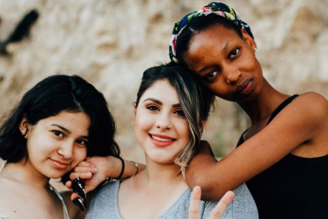 Portraiture of three diverse women leaning on each other's shoulders. The one in the middle is throwing up a peace sign.