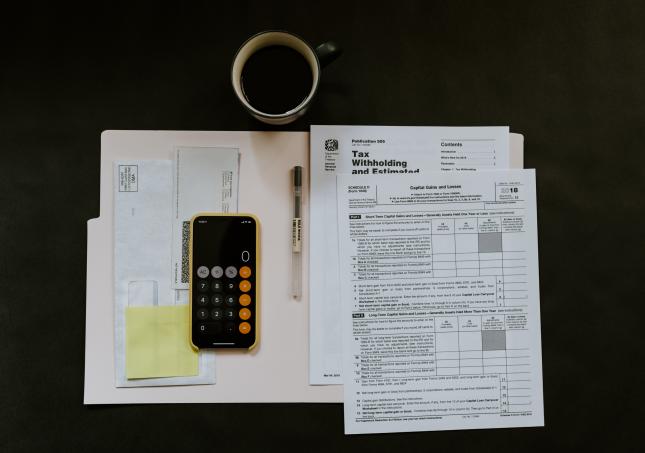 Overhead view of a manila folder containing tax documents, envelopes, and a calculator on top of a black desk next to a coffee mug